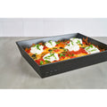 Ein schwarzes rechteckiges Ooni Detroit-Style Pizzablech 32 x 24 cm, gefüllt mit einem frisch gebackenen Gericht, belegt mit einer großzügigen Menge geschmolzenem Käse, Ricotta-Klecksen, schwarzen Oliven, Tomatenscheiben und gehackten grünen Kräutern. Die Pfanne steht auf einer grauen Arbeitsplatte mit einer weiß gefliesten Wand im Hintergrund.