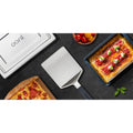 Auf einer dunklen Arbeitsplatte liegen ein Ooni Pizza-Servierheber von Ooni mit Klinge aus rostfreiem Stahl, ein rechteckiges Blech mit Pfannenpizza und Belag, ein Metall-Ofendeckel mit Ooni-Logo, ein Holzbrett mit Pizzascheiben und Kirschtomaten.