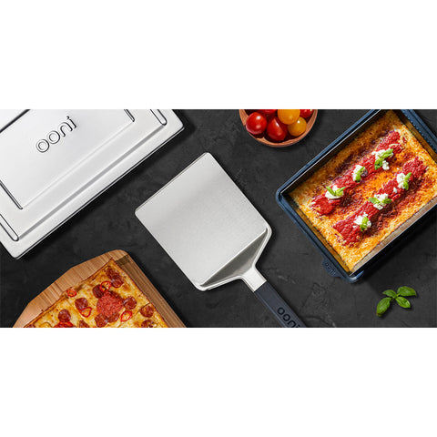 Auf einer dunklen Arbeitsplatte liegen ein Ooni Pizza-Servierheber von Ooni mit Klinge aus rostfreiem Stahl, ein rechteckiges Blech mit Pfannenpizza und Belag, ein Metall-Ofendeckel mit Ooni-Logo, ein Holzbrett mit Pizzascheiben und Kirschtomaten.