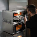 Ein Mann mit schwarzen Handschuhen zieht in seiner modernen Küche eine Pizza aus einem Edelstahl-Tischbackofen, der auf dem Effeuno Ofen Stapel-Kit, einer praktischen Stapelhilfe von Effeuno, steht.
