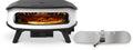 Ein moderner Cozze 17 Zoll Pizzaofen, Gas 50mbar mit rotierendem Pizzastein (CH-Regler) backt eine Pizza bei sichtbaren Flammen. Daneben liegt ein abnehmbares Metall-Frontpanel mit "cozze"-Schriftzug und Griff für perfekte Ergebnisse.