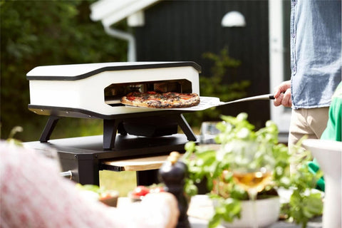 Eine Person benutzt einen Pizzaschieber aus Metall, um eine belegte Pizza aus dem Cozze 13 inch Pizzaofen, elektrisch V230 - CH-Stecker von Cozze zu nehmen. Im Vordergrund ist ein Tisch mit Kräutern, einem Getränk und verschiedenen Lebensmitteln zu sehen.