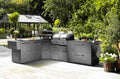Eine moderne Außenküche mit schlichten grauen Schränken, Gasgrill, Pizzaofen und Vorbereitungszone mit Cozze Elements Abfalleimer von Cozze auf einer Holzterrasse, umgeben von Grünpflanzen und einem Glasgewächshaus im Hintergrund.