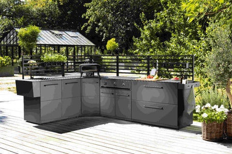 Eine stilvolle Outdoor-Küche mit dunklen Schränken, Grill und Spüle steht auf einer Holzterrasse mit der Cozze Elements Rückwand für Outdoor-Küchenmodul [110 cm] von Cozze, umgeben von üppigem Grün und einem Gewächshaus im Hintergrund.