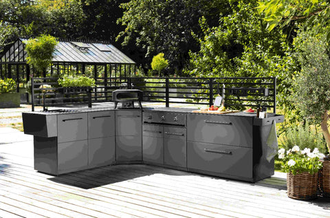 Eine moderne Outdoor-Küche mit schlichten grauen Schränken, Grill, Spüle und Arbeitsplatte auf einer Holzterrasse, dazu die Cozze Elements Rückwand für Outdoor-Küchen Eckmodul [90 cm], umgeben von Grün und einem Gewächshaus im Hintergrund.