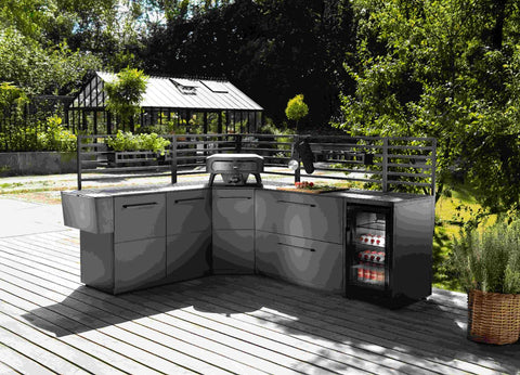 Eine moderne Outdoor-Küche mit grauen Schränken, Grill, Spüle und Getränkekühlschrank steht auf einer Holzterrasse. Dahinter steht die elegante Cozze Elements Rückwand für Outdoor-Küchenmodul [52 cm] von Cozze, mit Blick auf üppiges Grün und ein Glasgewächshaus.