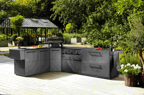Eine moderne Außenküche mit eleganten schwarzen Schränken, Grill, Pizzaofen, Spüle und der Cozze Toppingstation - schwarz von Cozze steht auf einer Holzterrasse inmitten von üppigem Grün mit einem Glasgewächshaus im Hintergrund.