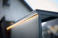 Nahaufnahme eines Cozze LED-Lichtstreifens für Elements Outdoor-Küchen [3m] von Cozze, der entlang einer Kante ein warmes Licht ausstrahlt, mit einem unscharfen Gebäude und einem schrägen Dach im Hintergrund.