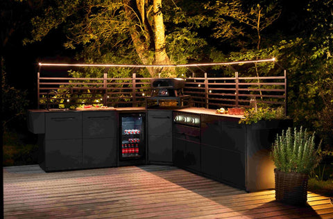 Moderne Outdoor-Küche mit schwarzen Schränken, Grill, Cozze Elements, kleinem Getränkekühlschrank und Speisen auf der Arbeitsplatte. Für stimmungsvolles Licht sorgen Cozze LED-Lichtstreifen [3m] zwischen Bäumen.