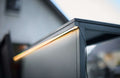 Eine Nahaufnahme einer modernen Cozze Elements Outdoor-Küche zeigt den Cozze LED-Lichtstreifen für Elements Outdoor-Küchen [5m], der am Rand entlang leuchtet, mit einem unscharfen Gebäude im Hintergrund.