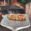 Eine glühend heiße Pizza mit goldenem Käse wird auf einem Metallschieber aus dem Burnhard Fat TONY Gas-Pizzaofen gehoben. Rauchschwaden steigen auf, während Pflanzen und eine Backsteinmauer die Kulisse bilden und die Qualität des Burnhard-Ofens unterstreichen.