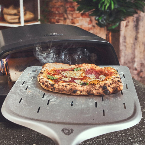 Eine glühend heiße Pizza mit goldenem Käse wird auf einem Metallschieber aus dem Burnhard Fat TONY Gas-Pizzaofen gehoben. Rauchschwaden steigen auf, während Pflanzen und eine Backsteinmauer die Kulisse bilden und die Qualität des Burnhard-Ofens unterstreichen.