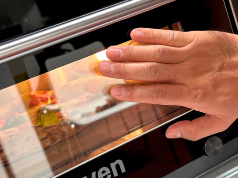 Eine Hand reinigt die Glastür eines Resto Italia Hooven 341 - Elektro Pizzaofen (Digital) mit einem Tuch oder Schwamm. Im Inneren wird Essen – möglicherweise eine leckere neapolitanische Pizza – zubereitet.