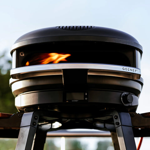 Vor einem verschwommenen natürlichen Hintergrund ist der Gozney Ofensims & Frontablage für Arc Outdoor-Pizzaofen auf einem stabilen Sockel montiert, mit sichtbaren Flammen im Inneren. Der Ofen mit seinem eleganten schwarz-silbernen Design, das für seine Hitzebeständigkeit bekannt ist, fällt deutlich auf.
