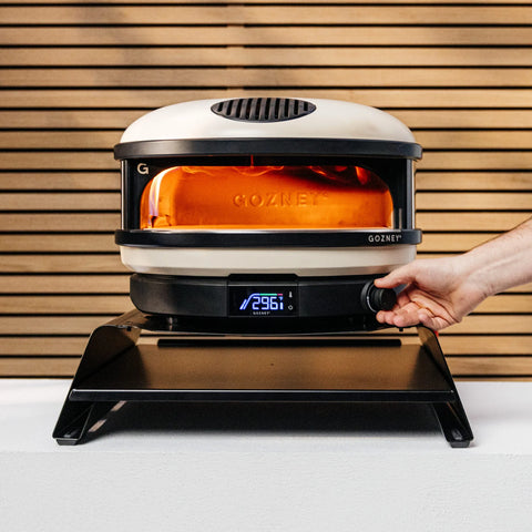 Eine Person stellt den Temperaturregler eines Gozney Arc Gas-Pizzaofens ein, dessen beleuchtete Innenseite das Gozney-Logo zeigt. Der Ofen steht auf einer Theke vor einem Hintergrund aus Holzlatten. Die Digitalanzeige zeigt eine Temperatur von 296°C an und verspricht Pizza in Restaurantqualität.