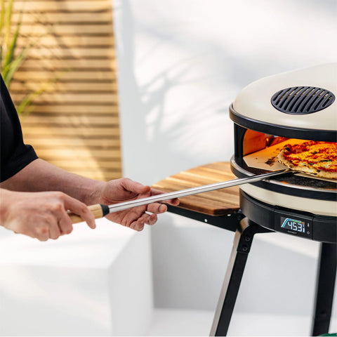 Eine Person verwendet einen runden Gozney Balance Pizzawender mit Buchenholzgriff und Aluminiumklinge, um eine fertig gebackene Pizza aus einem modernen Outdoor-Pizzaofen auf einem Dreibeinständer zu entnehmen. Die digitale Temperaturanzeige des Ofens zeigt 235 °C an und die Umgebung ist hell mit natürlichem Licht und minimalem Dekor.