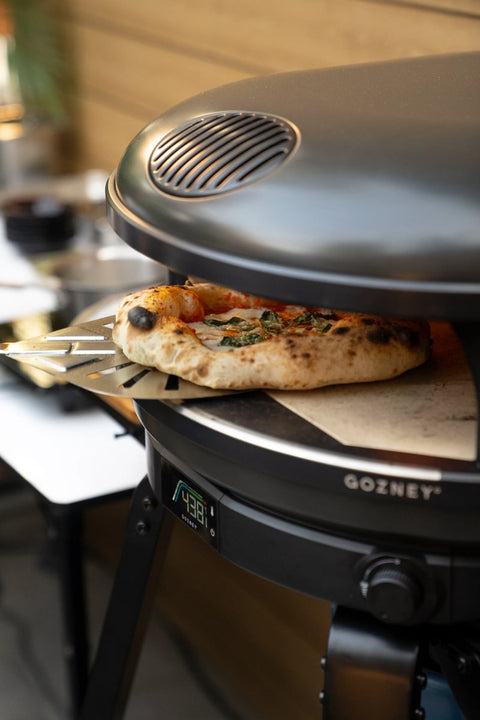 In einem Gozney Arc Gas Pizzaofen Off Black wird eine Pizza gebacken, die Temperatur auf dem Display zeigt 432 °C an. Die Pizza ruht auf einem Stein, wird von einer Schaufel gestützt und ist teilweise aus dem Ofen in einem einladenden Außenküchenbereich.
