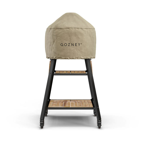 Ein abgedeckter Gozney Outdoor-Pizzaofen steht auf einem schwarzen Metallständer mit zwei Holzregalen. Die hellbraune Gozney Dome Abdeckhaube Kurz, mit dem Gozney-Logo versehen, sorgt mit ihren wetterfesten Materialien für Schutz. Der Ständer hat vier Räder für einfaches Bewegen.