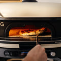 Eine Hand benutzt den Gozney Neapolitanischer Bogen für Dome XL (Gen 2), um eine knusprige, frische Pizza in und aus dem leuchtenden, modernen Gozney Dome XL zu schieben - ideal für authentisches neapolitanisches Pizzabacken.
