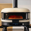 Ein moderner, cremefarbener Gozney Dome Gen 2 mit dem Gozney Neapolitanischer Bogen steht auf einem schwarzen Ständer. Seine Ofenöffnung leuchtet orange, ideal für neapolitanische Pizza. Holzlatten und eine weiße Wand bilden die Kulisse.