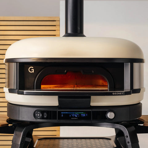 Ein moderner, cremefarbener Gozney Dome Gen 2 mit dem Gozney Neapolitanischer Bogen steht auf einem schwarzen Ständer. Seine Ofenöffnung leuchtet orange, ideal für neapolitanische Pizza. Holzlatten und eine weiße Wand bilden die Kulisse.
