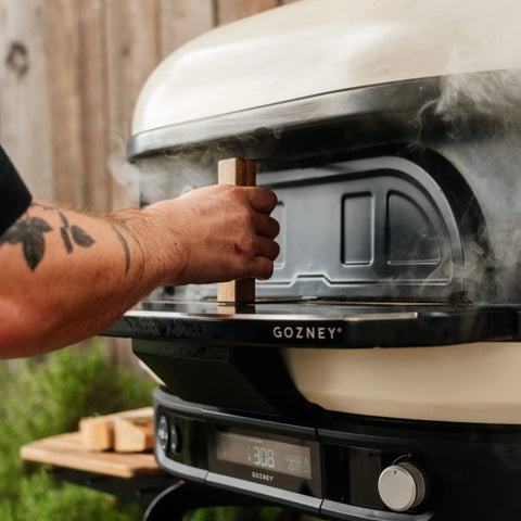 Eine Person mit einer Tätowierung am Arm öffnet mit einem Holzwerkzeug die Gozney Pizzaofentür für Dome XL (Gen 2) von Gozney an einem rauchenden Pizzaofen im Freien; daneben sind kleine Holzstücke und Grünpflanzen zu sehen.