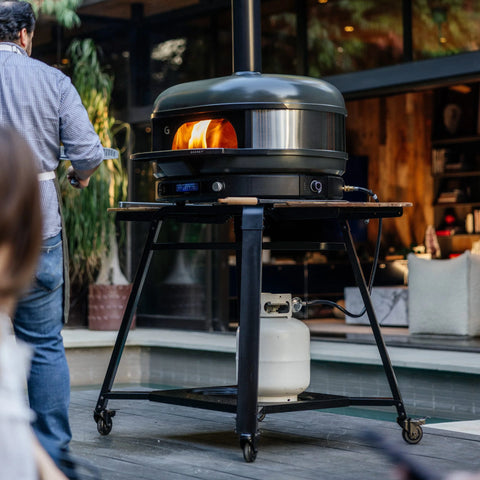 Ein Gozney Dome 2.0 Pizzaofen mit sichtbarer Flamme steht auf einem Gozney Stand & Pizzaofentisch für Dome 2.0 und Dome 2.0 XL von Gozney, mit einem Propantank darunter und einer Person, die in der Nähe einer modernen Terrasse steht, die zu einem gemütlichen Innenraum führt.