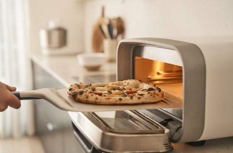 Eine Hand benutzt einen Pizzaschieber aus Metall, um eine frisch gebackene Pizza aus dem Ooni Volt 2 Elektro Pizzaofen & Halo Pro Spiral-Knetmaschine Bundle von Ooni in einer modernen Küche zu entnehmen.