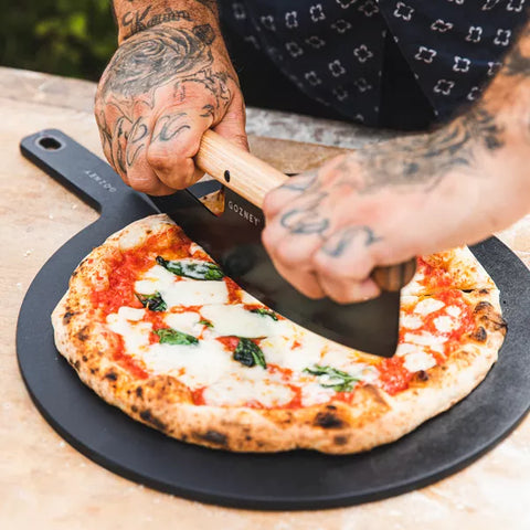 Eine tätowierte Person schneidet eine Pizza Margherita auf einem Gozney Pizza Servierteller (45 CM) von Gozney mit einem Rockerschneider mit Holzgriff und demonstriert dabei Stil und Präzision.