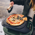 Jemand trägt eine grüne Jacke und schneidet eine dampfende Pizza mit Käse, Tomatensoße und Basilikum in einem Outdoor-Steinofen, zu dessen Ausstattung auch die Gozney Ablagefläche – Roof Rack for Tread gehört. Zum bequemen Schneiden der Pizza verwenden sie ein abnehmbares Schneidebrett.