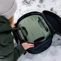 Eine Person in einer grünen Jacke und einer beigen Mütze legt ein mysteriöses grünes Objekt mit schwarzen Griffen in eine Gozney Venture Abdeckhaube & Tragetasche für Tread auf dem schneebedeckten Boden. Der Zweck des Objekts ist unklar, aber es scheint ein Behälter oder Gerät zu sein.