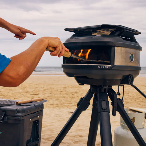 Auf einem Strandstativ steht ein tragbarer Pizzaofen, in den mit dem runden Pizzawender (15 cm) von Gozney Venture eine Pizza geschoben wird, während sich dahinter das Meer abzeichnet. Ein Propangastank und ein Getriebe stehen in der Nähe, und ein bewölkter Himmel unterstreicht den Charme des Meeres.