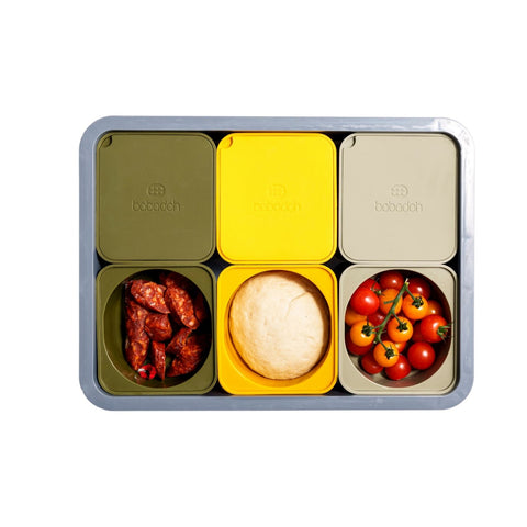 Eine Babadoh-Box – ein Stahlmetalltablett von Babadoh enthält sechs rechteckige Behälter: drei mit grünen und gelben Deckeln und drei offene, in denen Wurstscheiben, ein Brötchen und Kirschtomaten zu sehen sind.