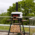 Ein moderner Gozney Dome Neapolitanischer Bogen und Schornsteinverlängerung von Gozney mit weißer Kuppel und schwarzem Sockel steht auf einer Terrasse. Darunter sind Holzscheite ordentlich auf Regalen gestapelt. Im Hintergrund sind ein üppiger grüner Baum und eine entfernte Stadtlandschaft zu sehen, die teilweise durch einen bewölkten Himmel sichtbar ist – perfekt für die Zubereitung authentischer neapolitanischer Pizza.