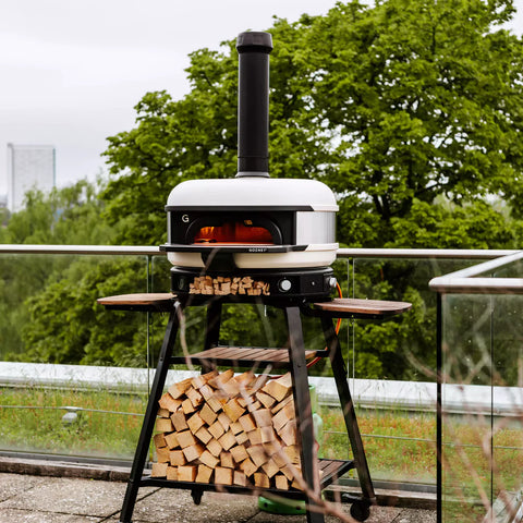 Ein moderner Gozney Dome Neapolitanischer Bogen und Schornsteinverlängerung von Gozney mit weißer Kuppel und schwarzem Sockel steht auf einer Terrasse. Darunter sind Holzscheite ordentlich auf Regalen gestapelt. Im Hintergrund sind ein üppiger grüner Baum und eine entfernte Stadtlandschaft zu sehen, die teilweise durch einen bewölkten Himmel sichtbar ist – perfekt für die Zubereitung authentischer neapolitanischer Pizza.