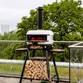 Ein moderner Pizzaofen für den Außenbereich auf einem schwarzen Ständer, gefüllt mit geschnittenem Brennholz. Der Ofen, ausgestattet mit der Gozney Dome Schornsteinverlängerung von Gozney zur verbesserten Luftzirkulation, hat einen Schornstein und ein aktives Feuer im Inneren. Er steht auf einem Dach mit einem Glasgeländer und bietet Aussicht auf grüne Bäume und die Skyline der Stadt in der Ferne.