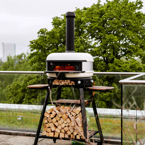Ein moderner Pizzaofen für den Außenbereich auf einem schwarzen Ständer, gefüllt mit geschnittenem Brennholz. Der Ofen, ausgestattet mit der Gozney Dome Schornsteinverlängerung von Gozney zur verbesserten Luftzirkulation, hat einen Schornstein und ein aktives Feuer im Inneren. Er steht auf einem Dach mit einem Glasgeländer und bietet Aussicht auf grüne Bäume und die Skyline der Stadt in der Ferne.