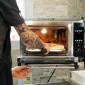 Eine Person mit tätowierten Armen bedient gekonnt einen Effeuno Pizzaofen P134HA PRO 509°C mit digitaler Steuerung, um eine Pizza aus dem Ofen zu nehmen. Der Ofen steht vor einer Wand aus hellen Steinfliesen, die dieser italienisch inspirierten Szene Eleganz verleiht.