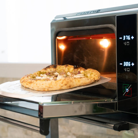 Vor einer Steinmauer wird eine Pizza mit goldbraunem Rand und Belag auf einer Schaufel aus dem glühenden Pizzaofen EffeUno P134HA PRO 509°C gezogen. Dieser in Italien hergestellte EffeUno-Ofen erreicht Temperaturen von 190°C und 150°C, während die Pizza auf der Arbeitsplatte abkühlt.