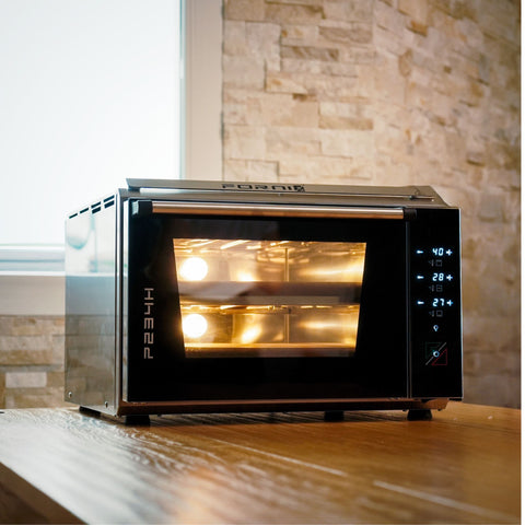 Der Effeuno Pizzaofen EffeUno P234H PRO 509°C ist ideal für perfekte neapolitanische Pizzen und besticht durch moderne digitale Bedienelemente und ein elegantes Edelstahl-Finish. Er steht elegant auf einer Holzoberfläche und das warme Licht seiner Innenbeleuchtung harmoniert perfekt mit einer Steinfliesenwand und einem Fenster.