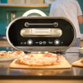 Der Evolve Smart Elektro Pizzaofen von Revolve steht auf der Küchenarbeitsplatte. Mit digitaler Anzeige für Zeit und Temperatur ist er von frisch gebackenen Pizzen auf Holzplatten umgeben.