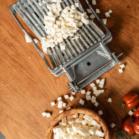 Ein Fumosa Profi Mozzarellaschneider mit Edelstahl-Schneidedrähte liegt auf einer Holzfläche und hat Weißkäsescheiben, von denen sich ein Teil in einer Holzschüssel befindet. Kirschtomaten an der Rispe liegen rechts neben dem Schneider.