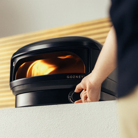 Eine Person stellt einen Drehknopf am schwarzen Gozney Arc Lite Gas Pizzaofen von Gozney ein, mit sichtbaren Flammen im Inneren, positioniert auf einer weißen Fläche vor einem Hintergrund aus Holzlatten.