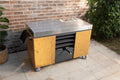 Die KILN Pizzastation von Everdure, ein hölzerner Outdoor-Küchenwagen mit einer glänzenden Edelstahl-Arbeitsfläche, schmückt die Terrasse. Sie verfügt über ein schwarzes Handtuch, mehrere Schubladen und ist von Topfpflanzen umgeben. Der Charme wird durch die Backsteinmauer und die grüne Kulisse noch verstärkt.