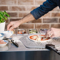 Eine Person verwendet die Everdure KILN Pizzastation, um eine Pizza zuzubereiten und verteilt Käse über Tomatensoße. In der Nähe stehen Schüsseln mit Aufschnitt und Gemüse sowie Behälter mit Belag auf einer Edelstahl-Arbeitsfläche neben einer Basilikumpflanze vor einem Backsteinhintergrund.