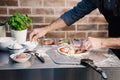 Eine Person verwendet die Everdure KILN Pizzastation, um eine Pizza zuzubereiten und verteilt Käse über Tomatensoße. In der Nähe stehen Schüsseln mit Aufschnitt und Gemüse sowie Behälter mit Belag auf einer Edelstahl-Arbeitsfläche neben einer Basilikumpflanze vor einem Backsteinhintergrund.