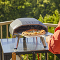 Jemand nimmt eine heiße Pizza aus dem Ooni Koda 12 by Ooni auf der Terrasse, umgeben von üppigem Grün.