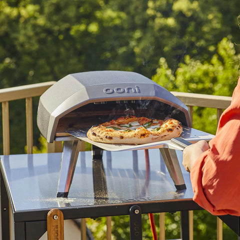 Jemand nimmt eine heiße Pizza aus dem Ooni Koda 12 by Ooni auf der Terrasse, umgeben von üppigem Grün.