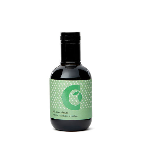 Eine kleine dunkle Glasflasche mit schwarzem Verschluss enthält Olivenöl extra vergine mit Basilikum-Aroma (100 ml). Die Flasche hat ein hellgrünes Etikett mit einem großen, stilisierten Buchstaben „C“ und einem Muster aus weißen Punkten, darunter der Text „G.B. Annunziati“, „Olio da condimento al basilico“ und der Markenname Ciccolella.