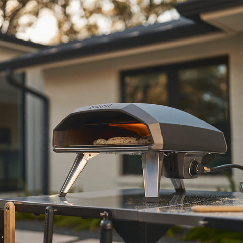 Der Ooni Koda 2 Gas-Pizzaofen von Ooni mit schlanken Metallbeinen und modernem Design steht auf einem Tisch und backt neapolitanische Pizzen im Hinterhof, im Hintergrund ist ein Haus zu sehen.